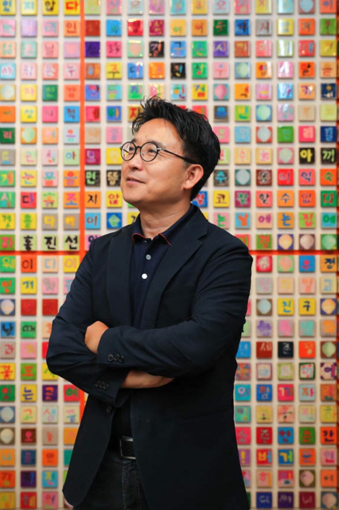 Ik-Joong Kang in front of a colorful wall of tiles in a black buttondown shirt